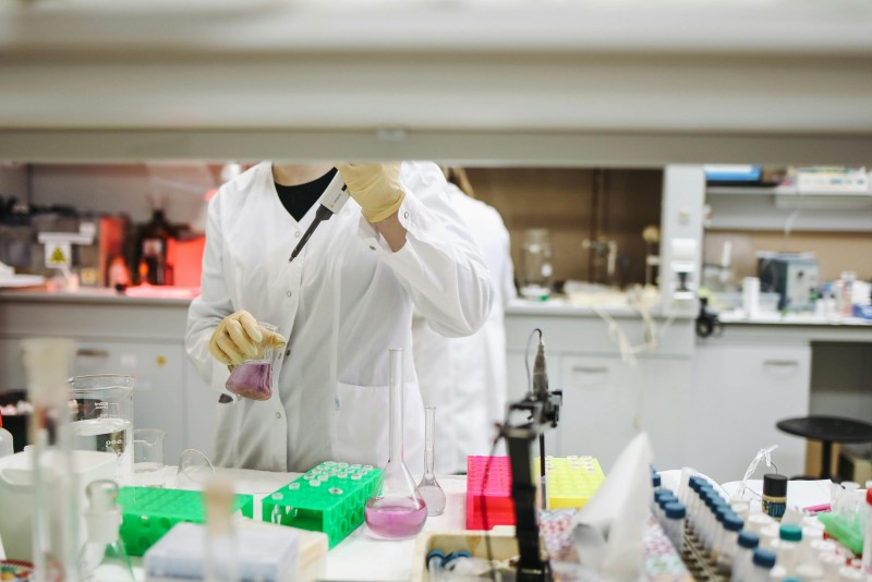 Entre estanterías de laboratorio se ve el tronco de un/a técnico trabajando con elementos de laboratorio