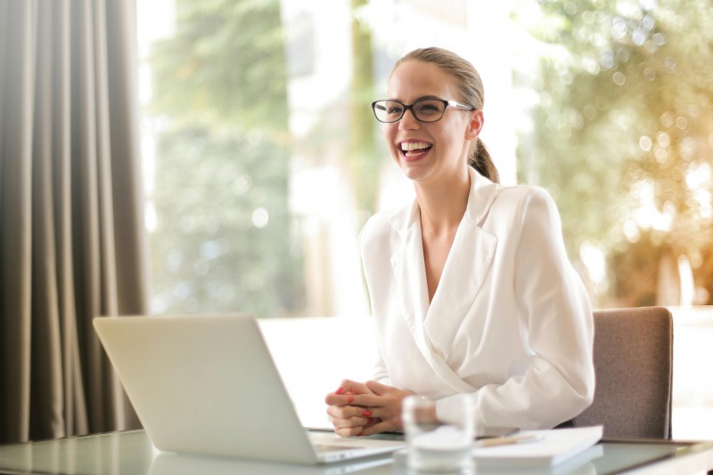 chica joven, castaña, con gafas negras y coleta, sonriendo en mesa de trabajo con ventanal de fondo donde se ven árboles  
