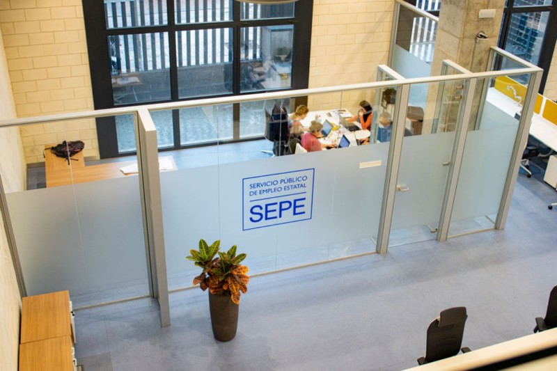 panorámica de una oficina del SEPE por dentro, con paredes de cristal esmerilado en blanco con logo y muebles en madera calra, una planta