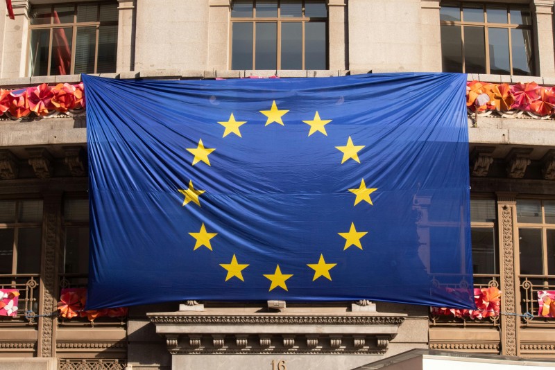 bandera de Europa estirada en una fachada con flores rosas y rojas al fondo